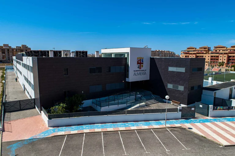 Modern British School of Almería building exterior with dark facade, school crest, and parking area under clear blue sky