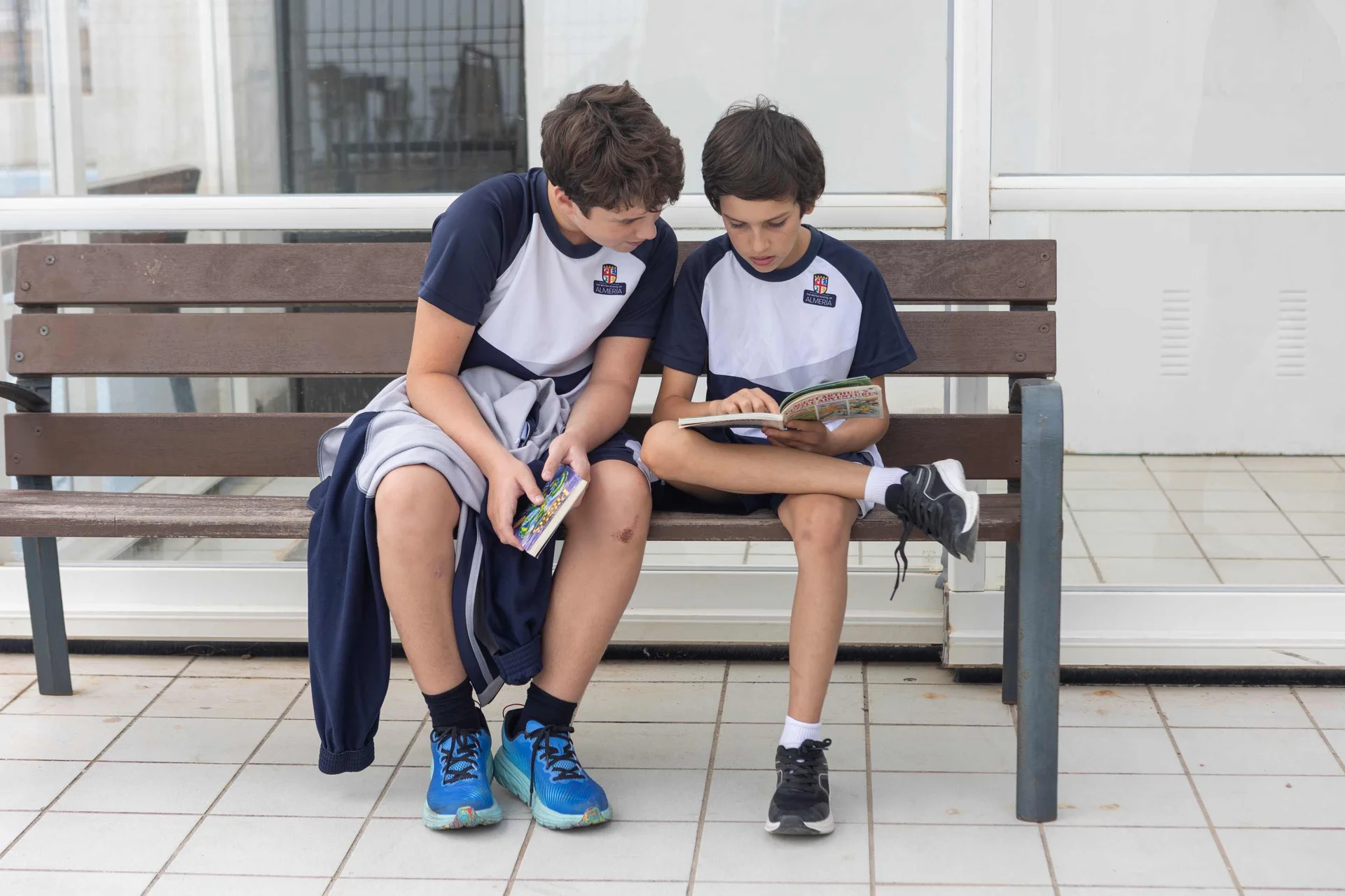 Two students in school sports uniforms sitting on a bench reading books together during break time