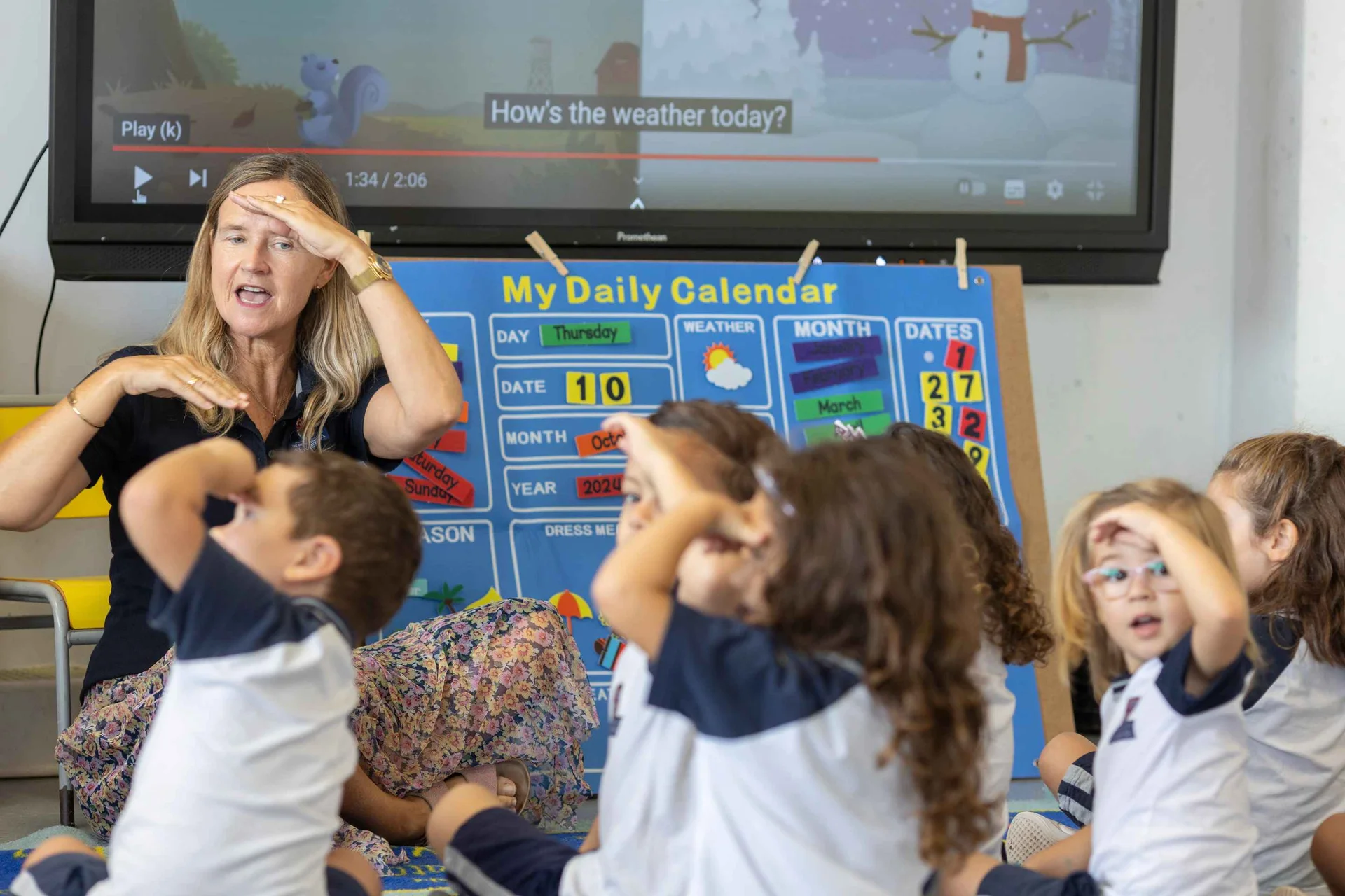 Teacher leading young students in a weather song activity, children mimicking hand-to-forehead gesture beside a daily calendar