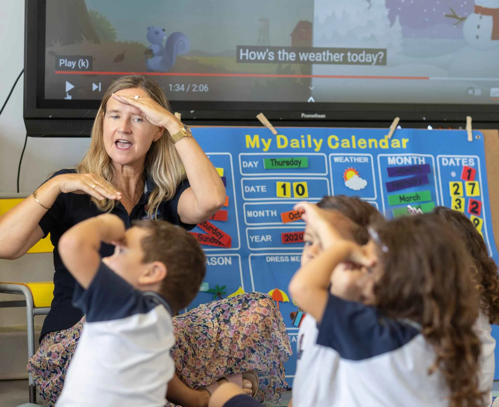 Teacher leading young students in a weather song activity, children mimicking hand-to-forehead gesture beside a daily calendar