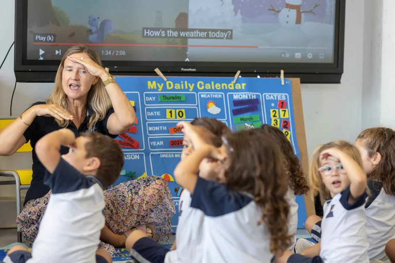 Teacher leading young students in a weather song activity, children mimicking hand-to-forehead gesture beside a daily calendar