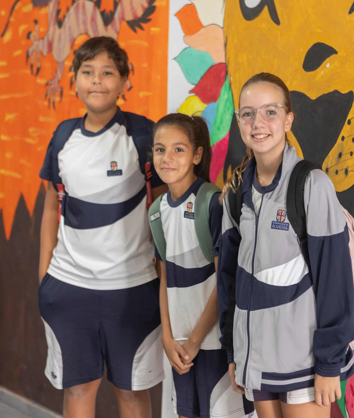 Three students in school uniforms smiling in a corridor decorated with colorful murals including a vibrant lion painting