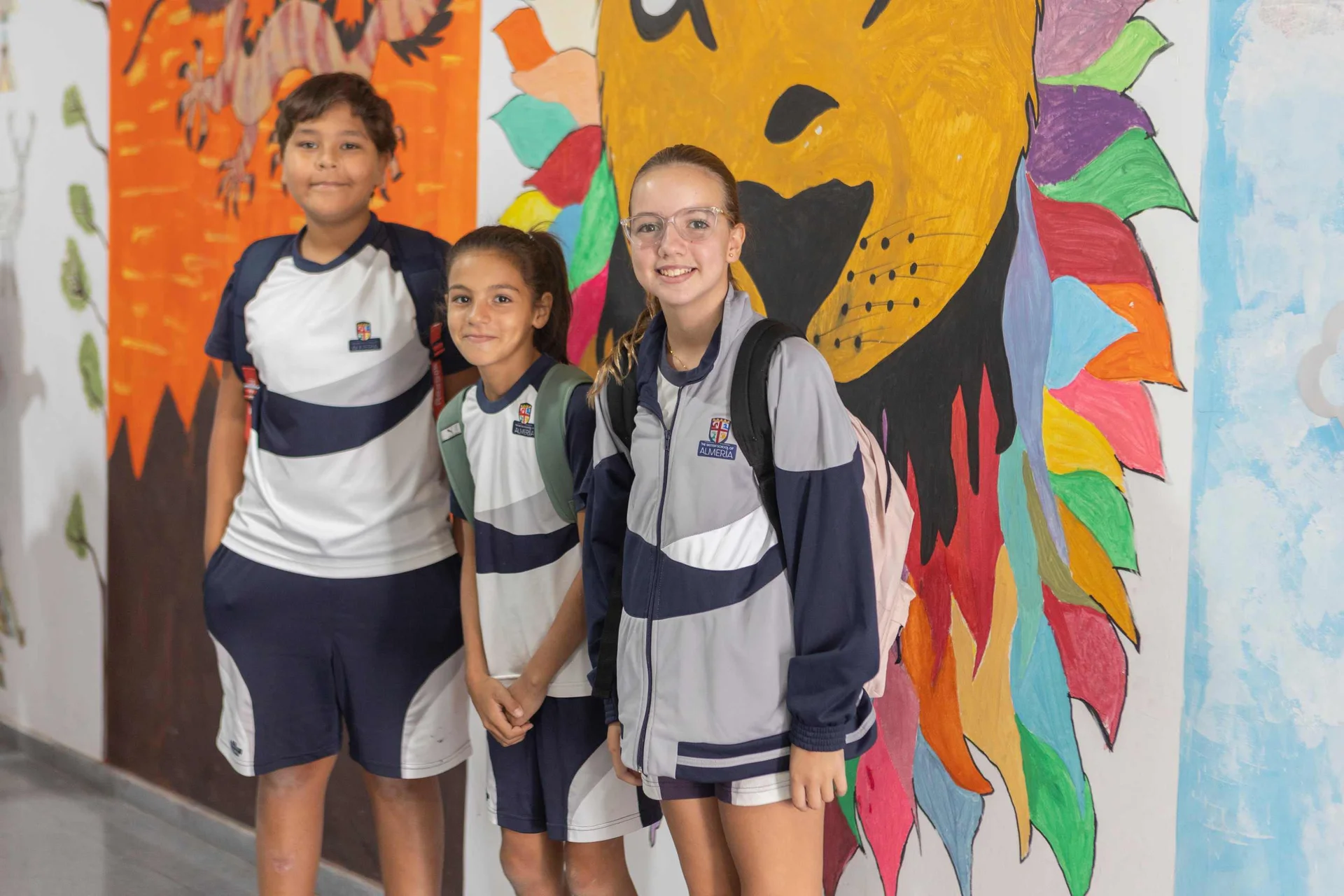 Three students in school uniforms smiling in a corridor decorated with colorful murals including a vibrant lion painting