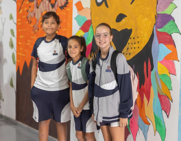 Three students in school uniforms smiling in a corridor decorated with colorful murals including a vibrant lion painting
