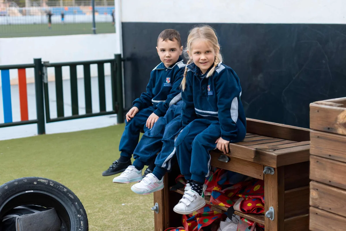 Two young students in navy blue school uniforms sitting on a wooden bench in the playground, smiling at the camera